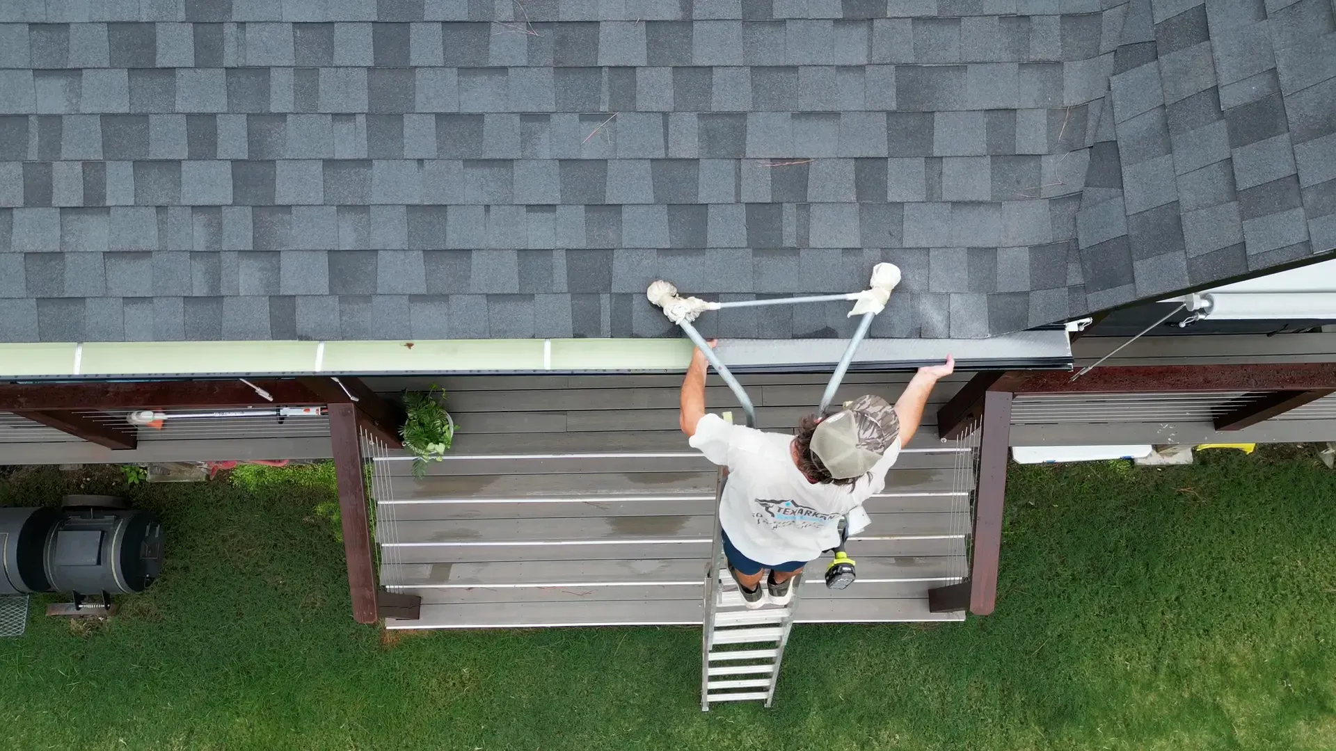 Overhead view of a technician working the gutter line along a gray shingle roof edge above a deck